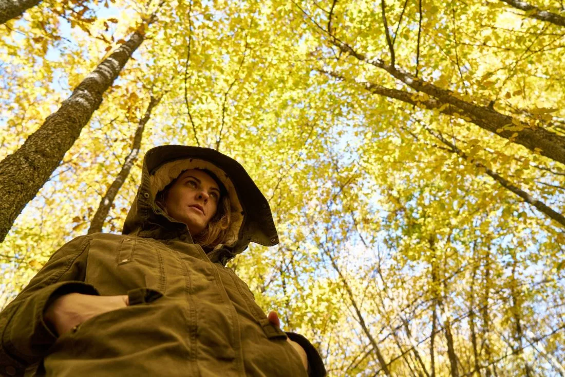 A woman in a hooded jacket standing under tall trees with bright yellow autumn leaves